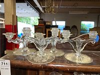 Four glass double candle holders placed on a wooden surface with visible background furniture, showing design and structural details from a front angle.