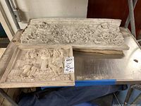 Both carved wall plaques placed on a metal surface showing size and condition including chips.