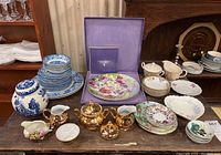 Mixed china plates, bowls, decorative plate in purple box, blue and white ginger jar, floral bowls and plates, gold-tone creamer and sugar set, and other serving pieces displayed on wooden table.