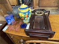 Photo of the lot including a double handled ginger jar with fruit motif, blue Disney metal tin, metal red toy cart, and an untested Bombay Co. wall clock on a wooden table.