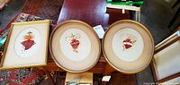 Three framed Sacred Heart needlepoint artworks displayed together, two oval frames and one rectangular frame, showing detailed and colorful embroidery designs.