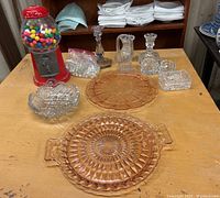 Lot on wooden table showing red carousel gumball machine filled with colorful gumballs, clear cut crystal glass candlestick holders, pitcher, containers, decorative glass marbles in bag, heart-shaped dish, and pink depression glass platters.