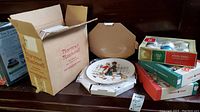 Photo showing Gorham Norman Rockwell collector plates in original packaging along with several boxes containing vintage glass Christmas ornaments.