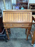 Front view of wood secretary desk showing vertical panel drop-front and single drawer with round knobs, turned legs, and wood grain finish.