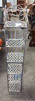 Full view of the tall, narrow metal shelf unit with four lattice shelves and decorative scrollwork on top and sides.