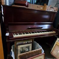 Steinway & Sons upright piano with closed fallboard and visible front side. Dark mahogany finish showing minor wear and scratches. Located in an indoor area crowded with other items.
