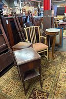 Photo displaying vintage furniture items including the marble top burled wood drum table, rattan armchair with themed seat upholstery, and two wooden side tables set on patterned rugs in a warehouse setting.