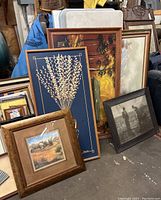 Photo showing multiple framed artworks leaning in a cluster including floral, landscape, abstract, and photo pieces