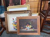 Five framed wall art pieces stacked and leaning against furniture showing various frame styles and artwork including floral and still life themes