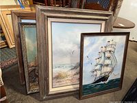 Four framed artworks stacked leaning against furniture. Mostly visible are two artworks: a tall sailing ship with white sails and a beach scene with ocean and seagulls.