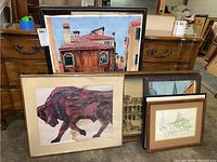 Five framed art pieces leaning against furniture, showcasing a variety of architectural and nature prints, including a prominently water-stained bull art print in the front.