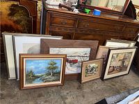 Nine framed paintings and drawings arranged leaning against wooden antique furniture, includes landscapes and still life compositions in various frame styles.