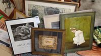 Photo showing multiple framed art prints and paintings stacked together, including a visible Ansel Adams print and a painting of a young girl in white dress.
