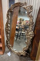 Full view of the ornate wall mirror with gold-tone decorative plastic frame featuring floral and scroll accents, leaning against a wooden panel in a storage setting.