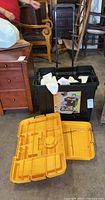 Black plastic work box tote with pull handle containing white socks and a detached yellow plastic box lid or organizer in foreground, surrounded by wooden furniture.
