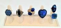 Front view of all six blue stone cocktail rings displayed on white ring stands, showing different shapes and styles of vintage rings.