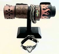 Six vintage leather and faux leather bracelets displayed on a black T-bar stand, plus one separate black faux leather bracelet with silver heart embellishments.