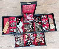 Open black vintage jewelry box showing top trays filled with assorted vintage costume jewelry items including chains, brooches, earrings, and pins.