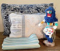 Overview showing two throw pillows, folded striped fabric, and a blue feathered Kachina doll standing on a wooden base.
