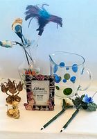 Photo showing two glass pitchers, decorative picture frame with butterfly accents, two metal candle holders, and novelty pens with flower and feather decorations arranged on a white surface