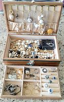 Overall view of open wooden jewelry box filled with assorted vintage jewelry including necklaces, rings, bracelets, and earrings arranged in compartments and hanging from lid hooks.