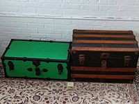 Two antique flat top steamer trunks resting side by side on a patterned rug against a white brick wall. One painted green with black hardware, the other wood with black panels and metal features.