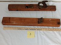 Top view of two wooden carpenter planes, one with handle and one without, placed next to a 36-inch ruler for scale. The wood shows age and use marks.