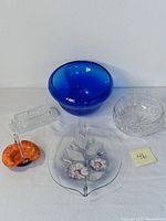 Front view of blue centerpiece bowl, clear glass tray with flower accents, clear candy dish, rectangular butter dish, ashtray, and small decorative glass basket with orange and yellow colors and clear handle.