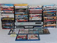 Photo showing a wide view of two DVD players with a large collection of DVDs and Blu-rays stacked behind them and some in front.