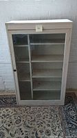 Front view of the white mid-century cabinet with a glass door showing 7 shelves inside.