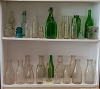 Full shelf view showing two rows of assorted vintage glass bottles, some clear, green, and blue, including medicine, dairy, and beverage bottles.
