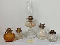 Five vintage pressed glass hurricane chimney lamps arranged in a row including one tall lamp with glass chimney, two clear glass lamps with rounded bases, one clear horizontal ribbed glass lamp and one small amber glass lamp.