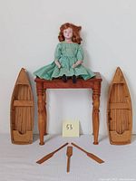 Marion doll seated on the small wooden doll house table with two wooden model rowboats on either side and three wooden oars in front on the floor.