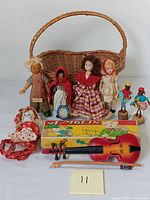 Full view of all dolls, wicker basket, toy violin, and box arranged on a white surface with a numbered label '11'.