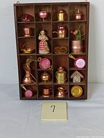 Full view of wooden shadow box containing various miniature copper cookware and decorative figures arranged in compartments.