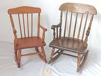 Two vintage wooden children's rocking chairs side by side, one darker with stencil decoration, the other lighter brown, both with spindle back and armrests.