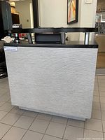 Front-left perspective of desk showing grey textured front panel and black glass shelf on top