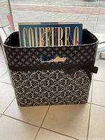 Magazine basket with visible hairstyling book titled 'COIFFURE 7' inside, showing front of basket with black and white floral and geometric patterns.