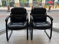 Front view of the pair of black vinyl office chairs with white stitching showing padded seats and backrests, designed with armrests, placed side by side.