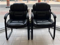 Front view of two black vinyl office chairs showing padded seats, backs, and armrests with signs of wear on upholstery.