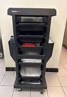 Front view of the Babyliss Pro rolling salon accessory tower showing multiple black trays and side compartments on caster wheels, with some wear visible.