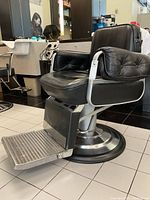 Front angle view of the vintage Belmont barber chair showing the black leather upholstery with signs of wear, thick padded armrests, and metal footrest.