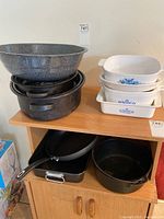 Stack of large enamel speckled roasting pans and black cast iron pots along with white ceramic casserole dishes with blue flower decoration.