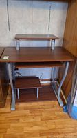 Front view of a metal and laminate computer desk showing the desktop, elevated small shelf, lower shelf, and metal legs on wood floor.