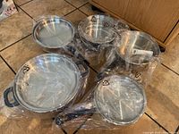 Photo showing six stainless steel cookware pieces sealed in plastic wrap with black handles and one steamer insert.