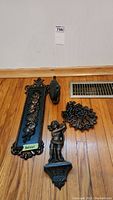 Three black metal decorative items on wood floor: ornate rectangular floral wall art panel, bird-shaped wall hook, and angel figurine.