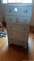 Front view of five drawer wooden dresser painted white with two round knobs on each drawer, decorative floral motifs on two drawers, showing wear.