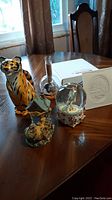Photo showing all four items on a wooden table with window background: tiger-striped vase, lithophane, music globe, and ceramic shoe bank.