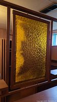 Large wooden entertainment wall unit with amber textured glass panel in the center. The glass panel is framed and mounted in the structure, showing a distinct textured pattern and amber tint. The wood frame is medium brown with visible wear.