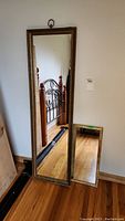 Full view of the two mirrors standing on the floor against a white wall, showing the taller wood framed mirror and smaller metal framed mirror angled on floor.